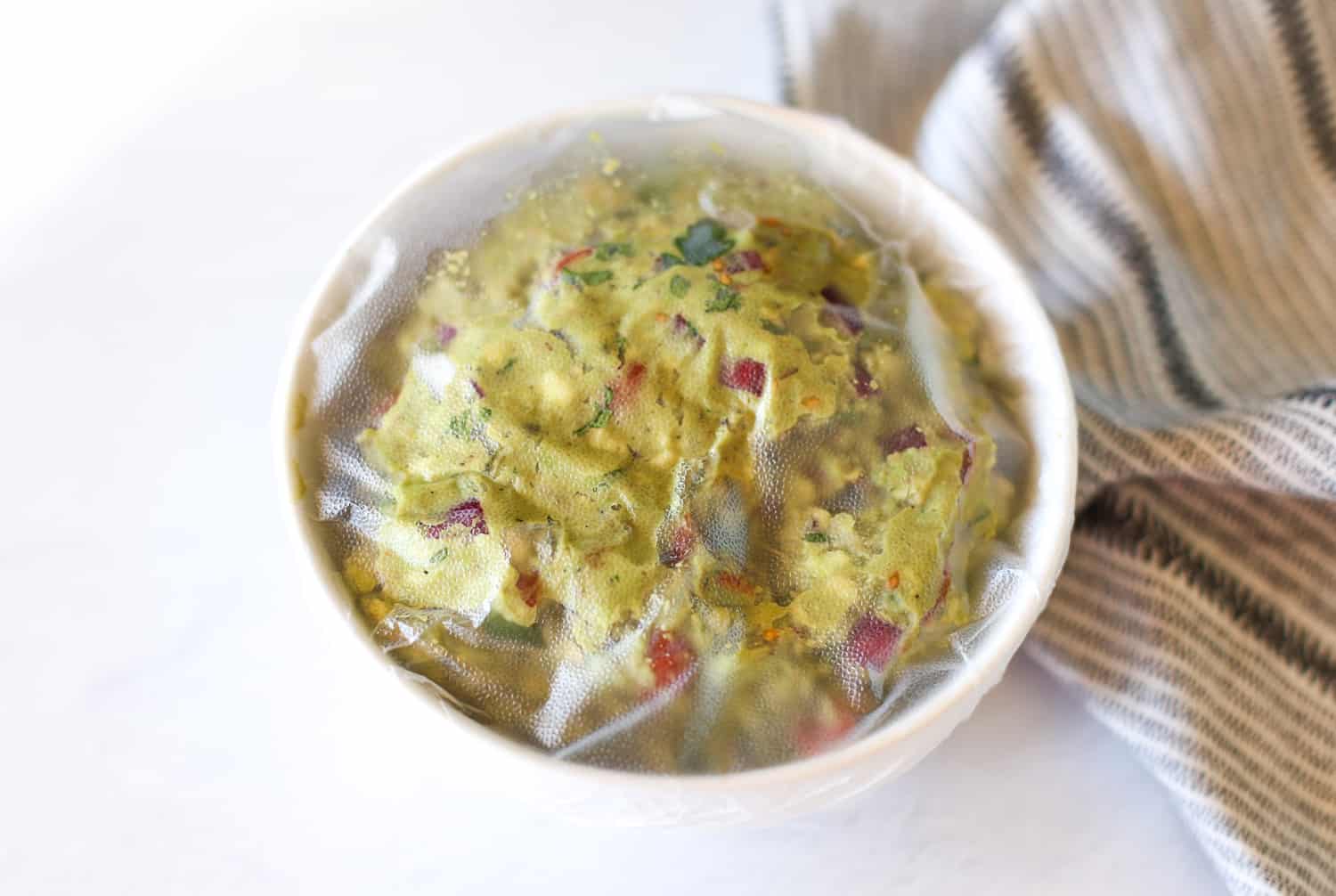 A bowl of homemade guacamole covered with plastic wrap.