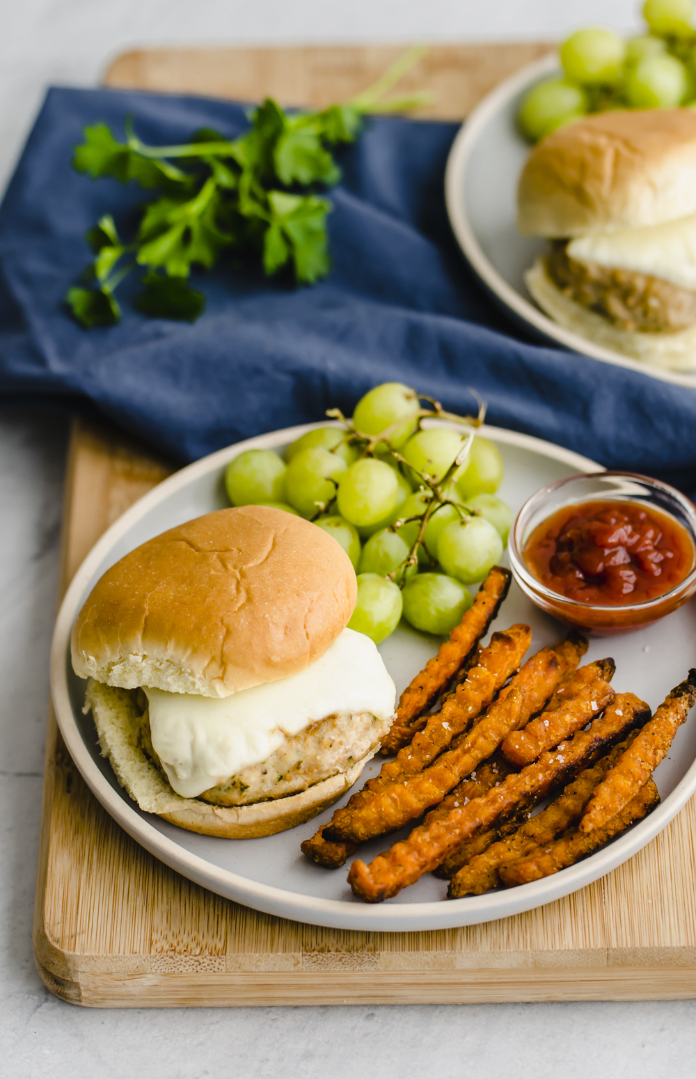 Mini chicken burgers on white plates with sweet potato fries, and grapes.