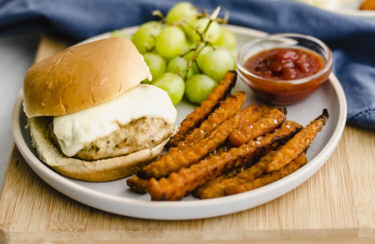 Chicken burger on a bun with cheese on a plate with sweet potato fries and grapes.