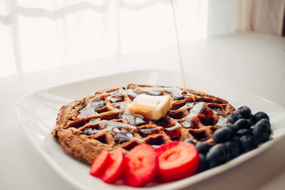 A waffle on a plate with a pat of butter on top and syrup being poured over it with strawberries and blueberries on the side.