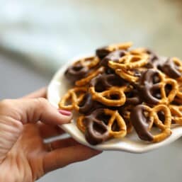 A hand holding a plate of dark chocolate and peanut butter covered pretzels.
