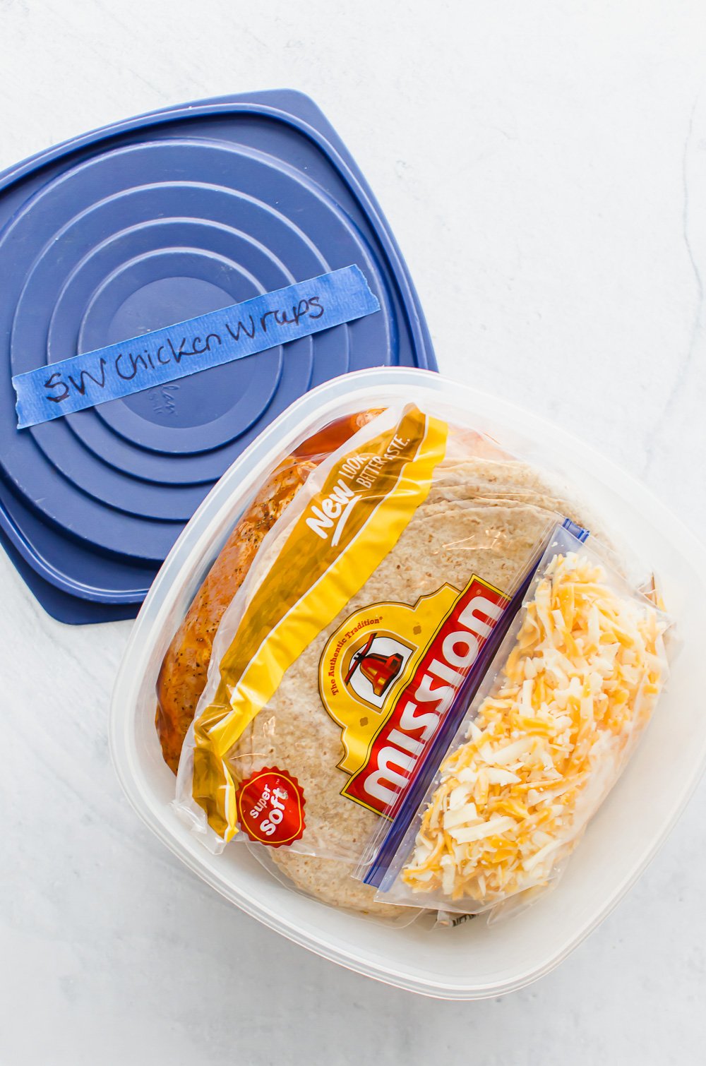 Southwest chicken wraps packaged as a freezer meal with baggies of cooked bacon, shredded cheese, marinating chicken, and tortillas in a square plastic container.