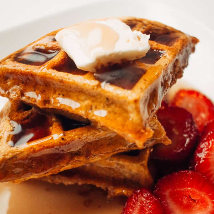 Stack of waffles on a plate with syrup and butter on top and strawberries on the side.