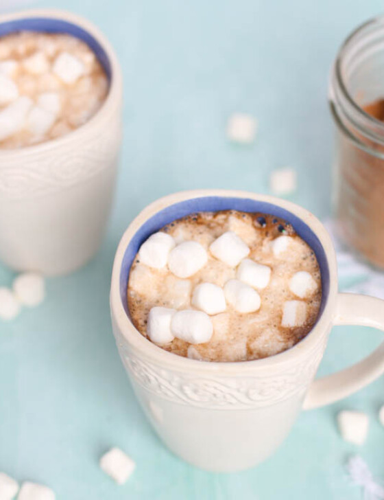 Two white mugs of homemade hot chocolate with mini marshmallows on top and sprinkled around the table.