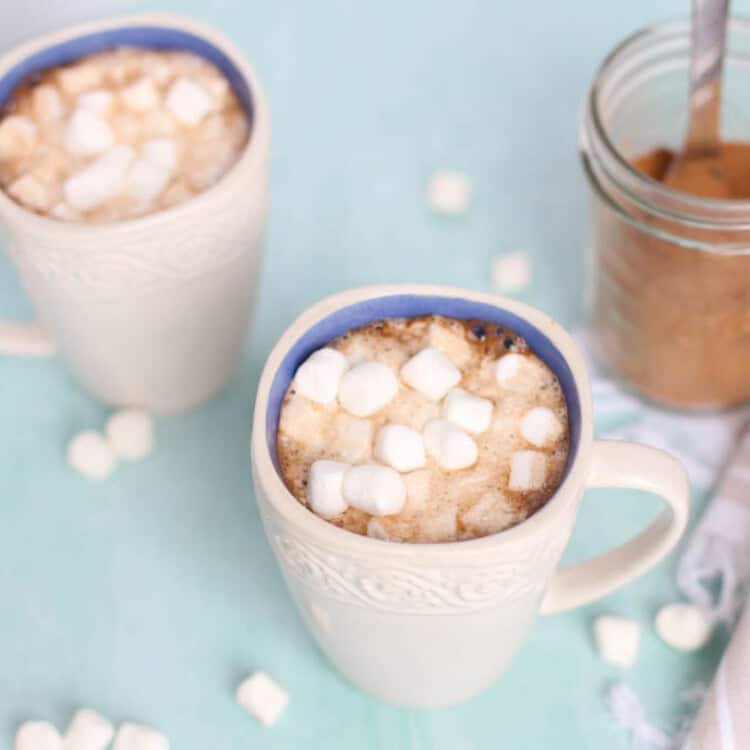 Two white mugs of homemade hot chocolate with mini marshmallows on top and sprinkled around the table.