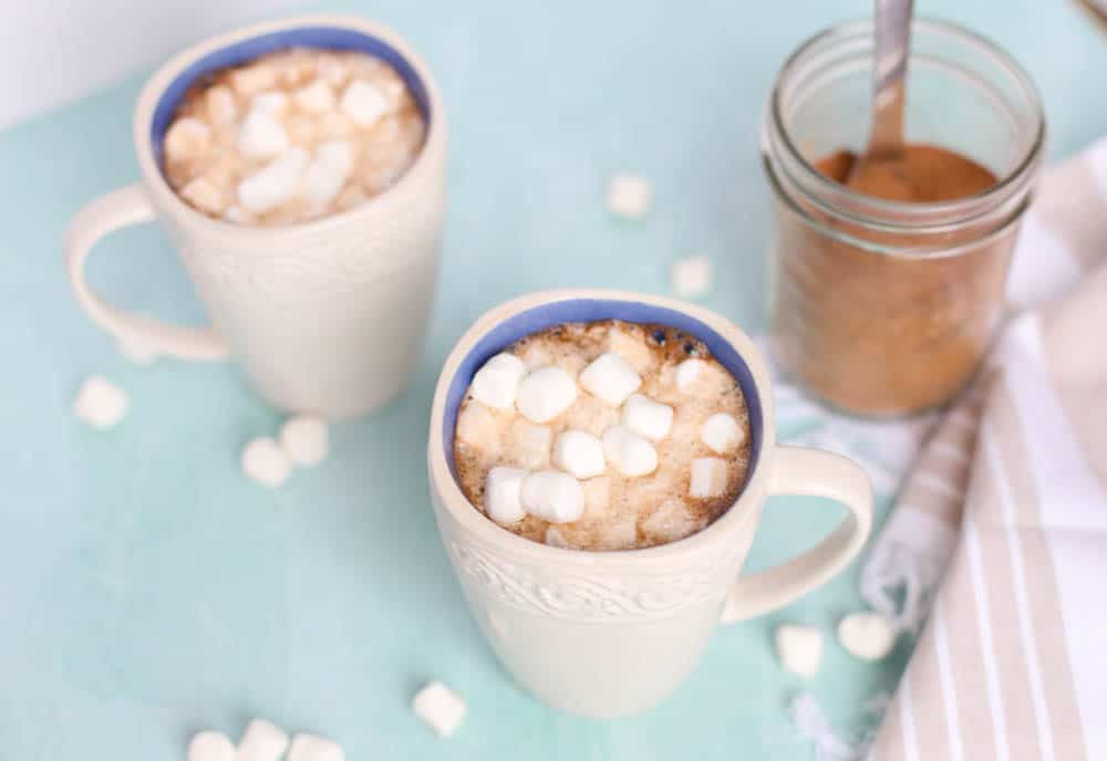 Two mugs of homemade hot chocolate with mini marshmallows on top and sprinkled on the table.