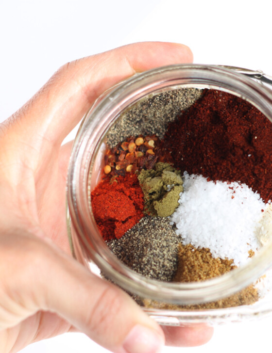 A hand holding a small jar with all of the individual spices that go into homemade taco seasoning.
