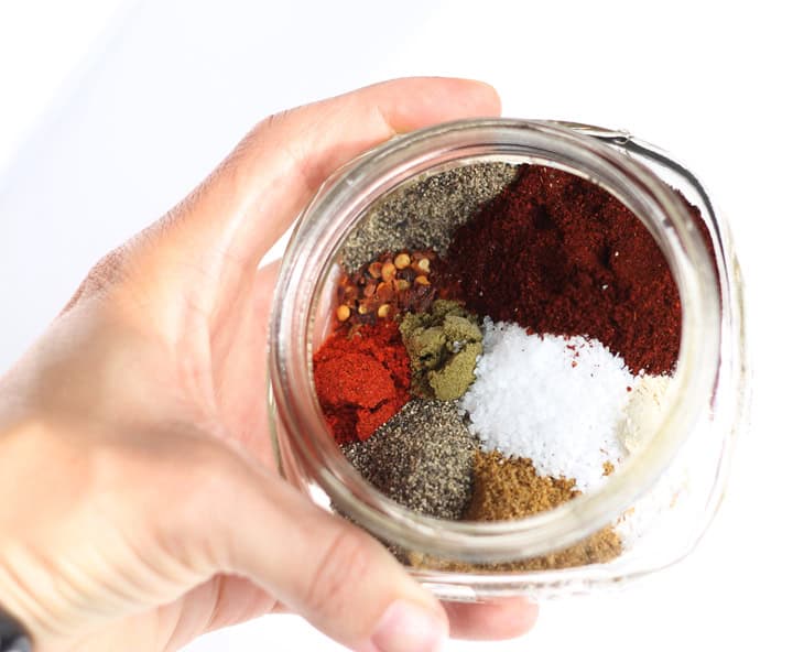 A hand holding a small jar with all of the individual spices that go into homemade taco seasoning.