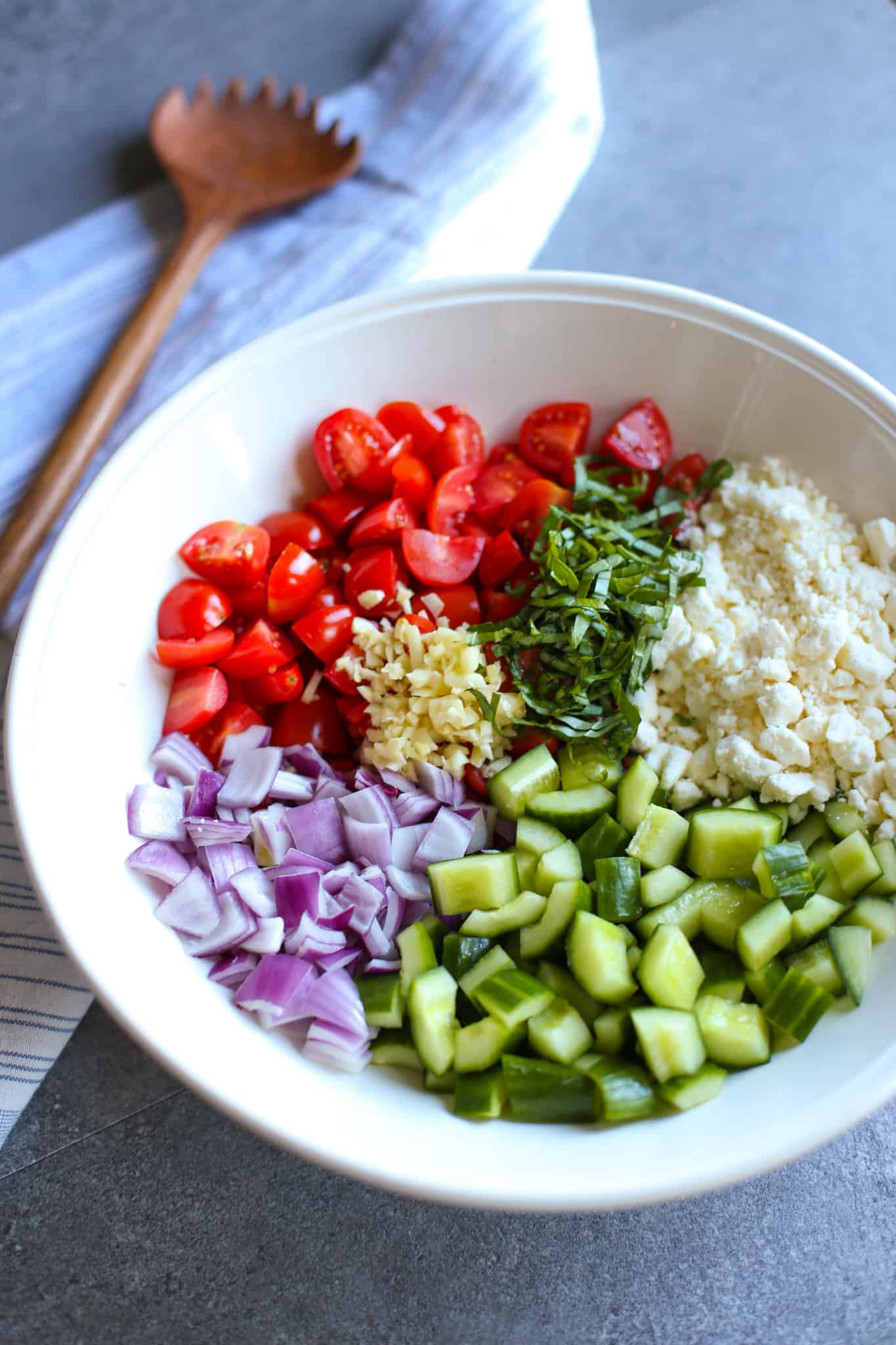 Chopped cucumber, red onion, tomatoes, basil, garlic and crumbled feta in a white bowl.