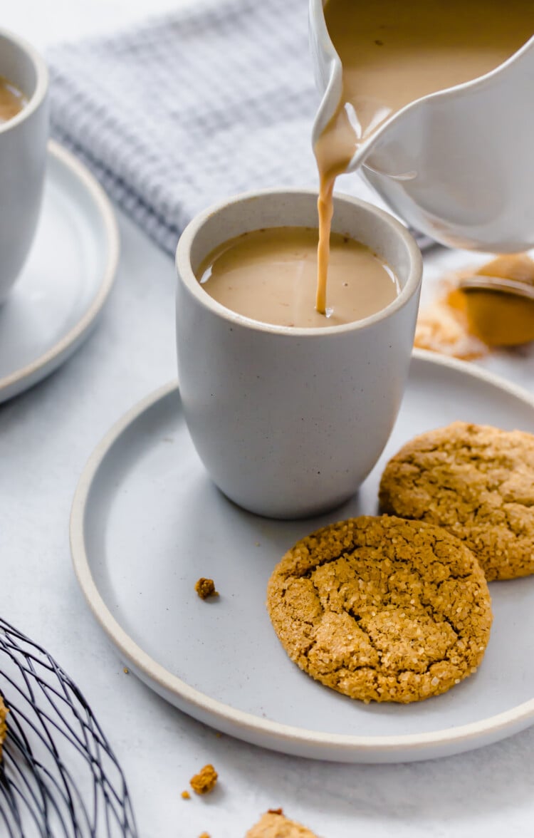 Pumpkin spice latte being poured into a white mug with cookies sitting on a plate next to it.