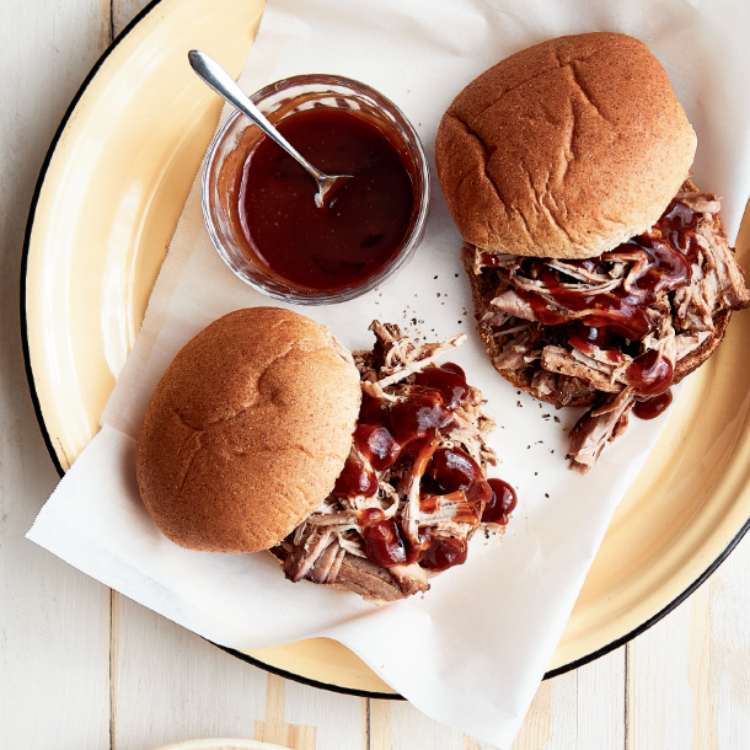 Two pulled pork sandwiches on a plate with a small bowl of BBQ sauce on it as well.