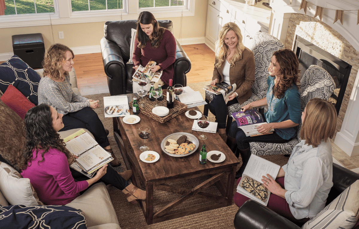 A freezer club of friends sitting around a living room with cookbooks.