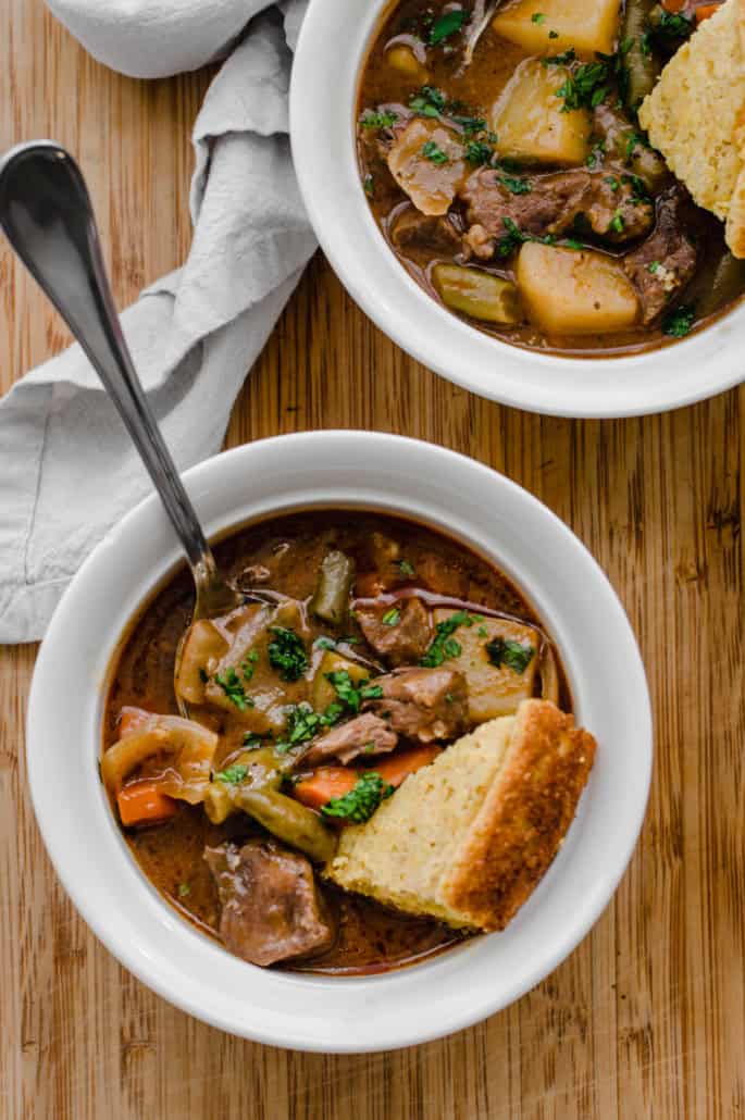 Bowls of easy beef stew with potatoes, carrots, onions, green beans, and stew meat in it. On a wooden table.