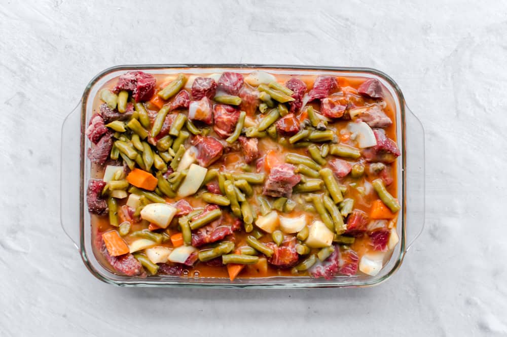 Uncooked beef stew ingredients in a casserole dish with stew meat, onions, carrots, green beans, and cream of mushroom soup.