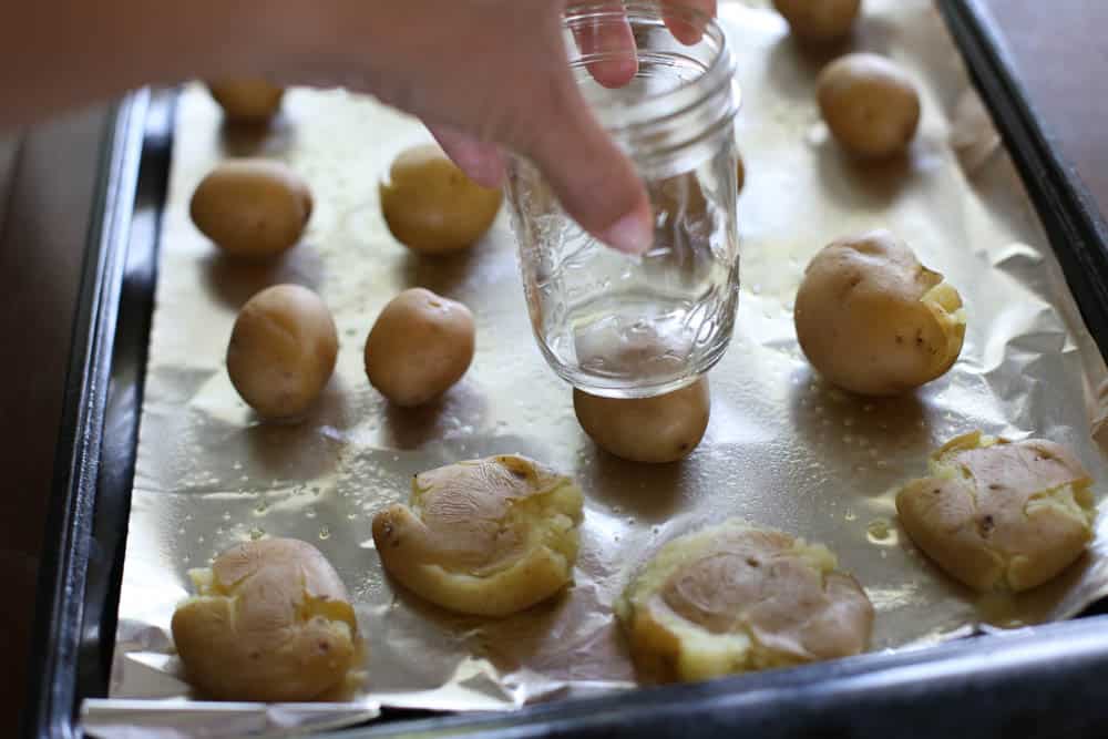 A mason jar being used to smash yukon potatoes on a foil-lined baking sheet.