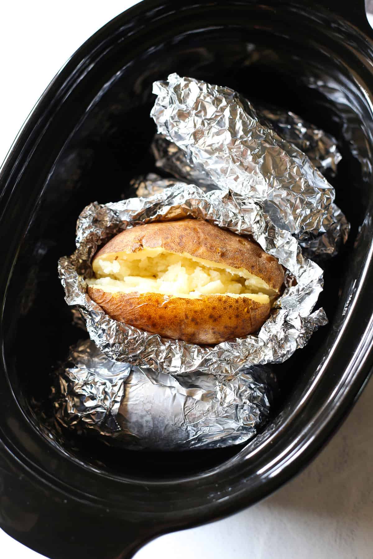 A cooked baked potato sitting on top of foil wrapped potatoes in a crock pot. 
