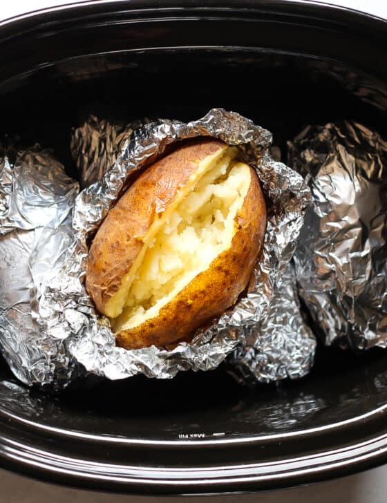 A cooked baked potato sitting on top of foil wrapped potatoes in a crock pot.