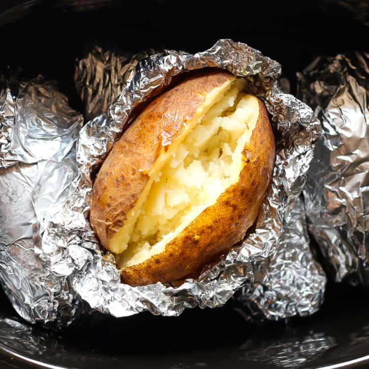 A fully cooked baked potato split open placed on top of other foil wrapped potatoes in a crock pot.