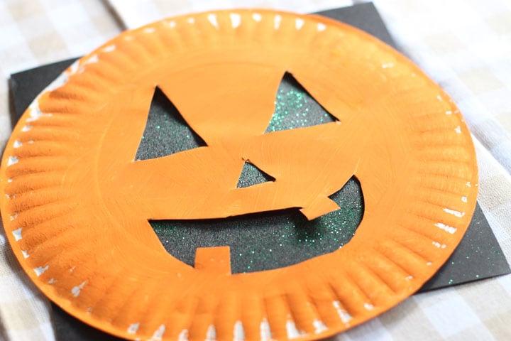 An orange-painted paper plate with a cut-out pumpkin face laying on top of a piece of black paper with glitter on it.