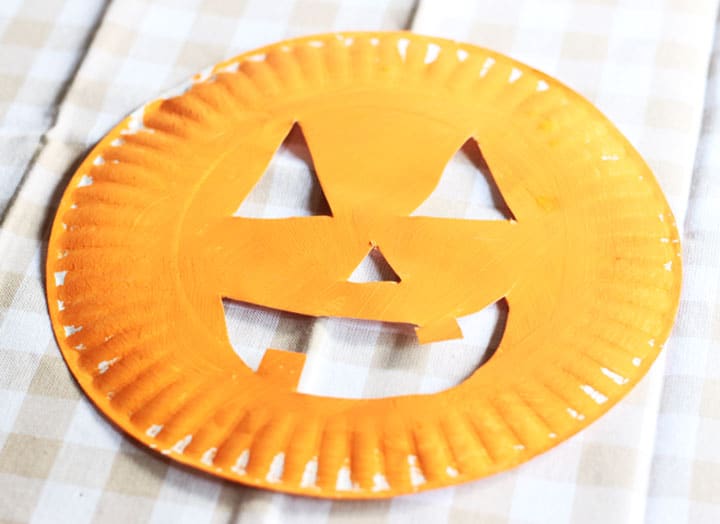 A paper plate that's been painted orange with a face cut out to look like a pumpkin. 