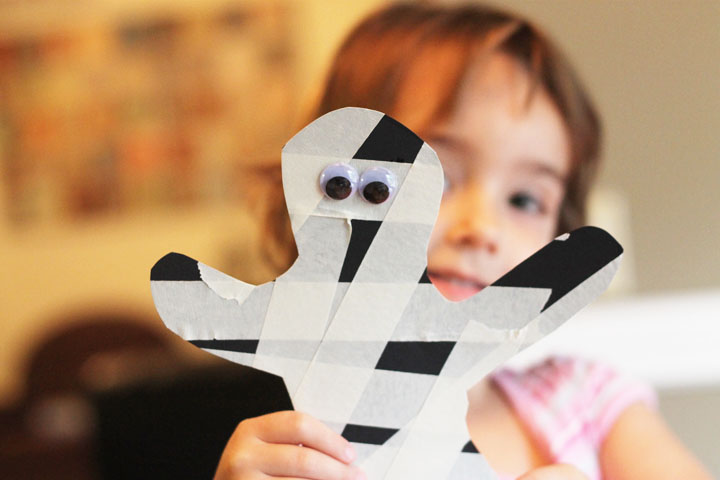 Child holding up a completed masking tape mummy craft - black construction paper in the shape of a cut out with masking tape put on to make it look like a mummy.