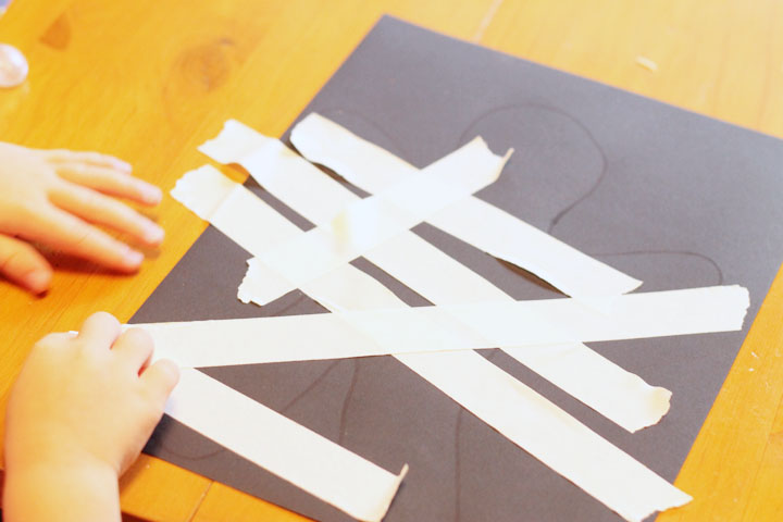 Black construction paper with an outline of a person on it and masking tape being put on the paper by child's hands.