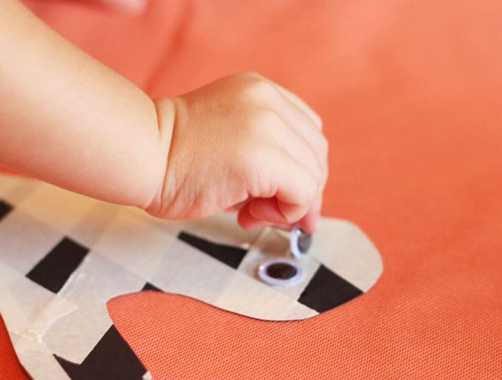 A child's hand placing a self-adhesive googly eye on a masking tape mummy craft.