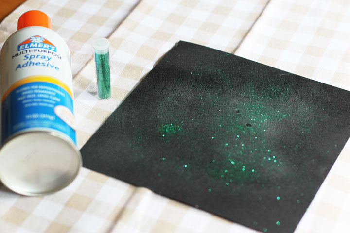 A black piece of paper with glitter on it with a can of spray adhesive and a small container of green glitter next to it.