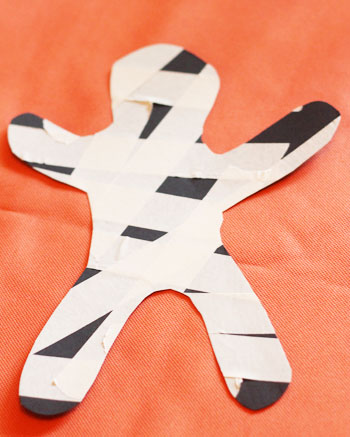 The shape of a person cut out of black construction paper, covered in masking tape and sitting on an orange piece of paper.