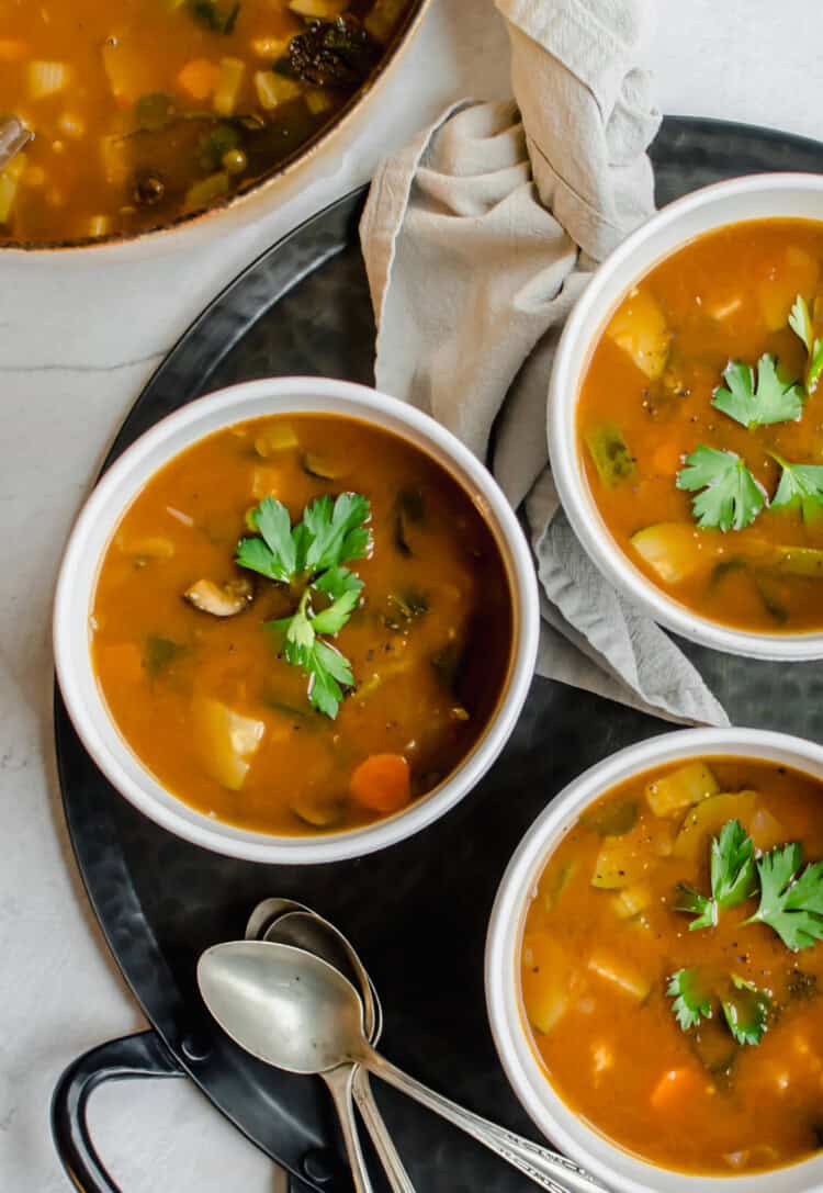 Large pot of vegetable soup with three bowls of vegetable soup. Spoons and napkins and fresh herbs on the side.