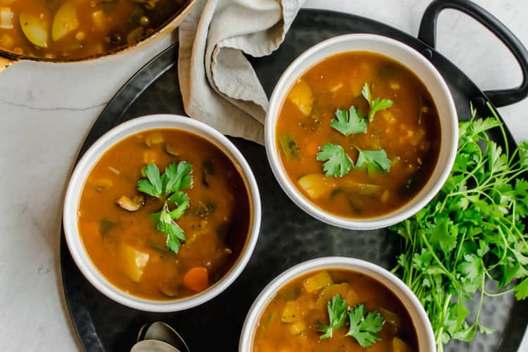 Large pot of vegetable soup with three bowls of vegetable soup. Spoons and napkins and fresh herbs on the side.