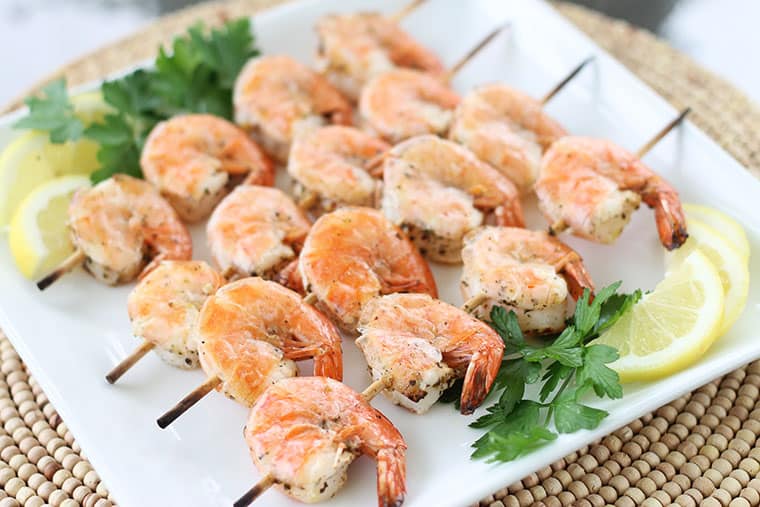 Four skewers of cooked Mediterranean shrimp on a white plate with lemon wedges and fresh parsley on the sides.