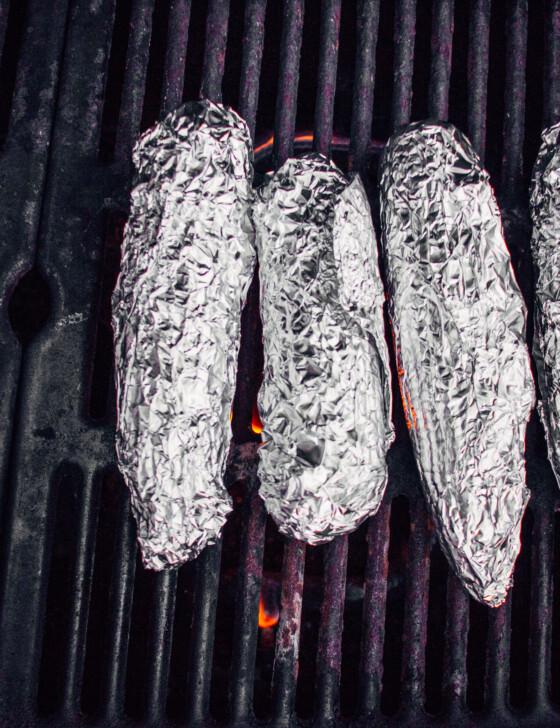 Four ears of corn on the cob wrapped in damp paper towels and then foil sitting on a grill with a flame underneath.