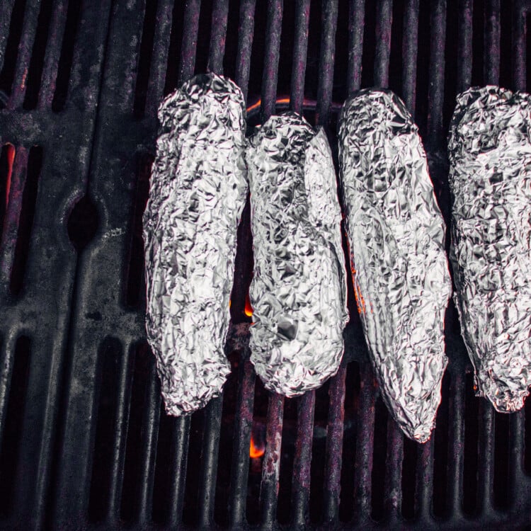 Four ears of corn on the cob wrapped in damp paper towels and then foil sitting on a grill with a flame underneath.