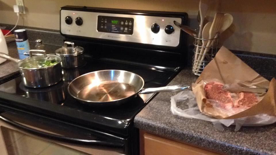Empty skillet on a stovetop with two raw steaks on the countertop next to it.
