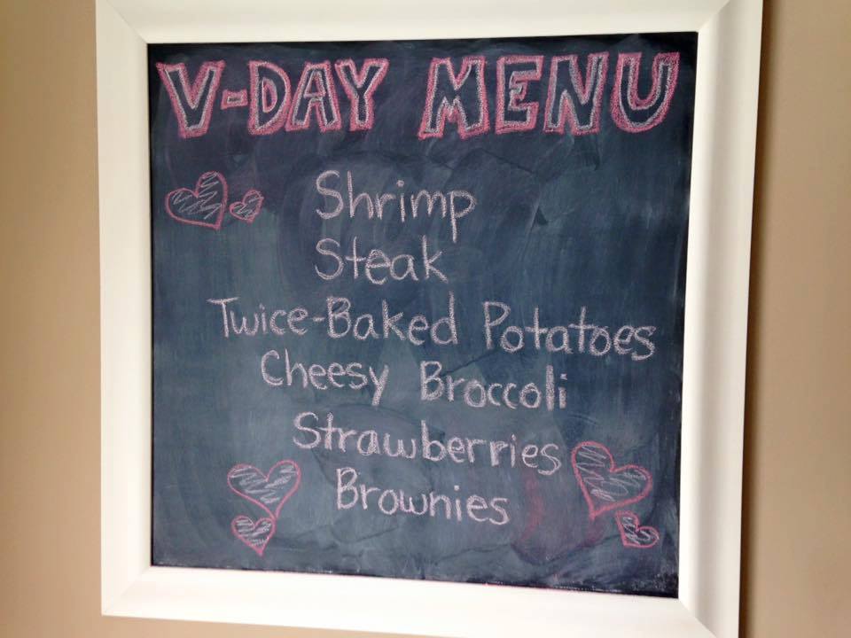 Valentine's day menu written on a chalkboard: Shrimp, Steak, Twice-Baked Potatoes, Cheesy Broccoli, Strawberries, Brownies.