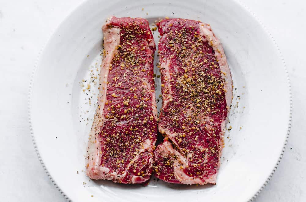 Two raw sirloin steaks on a white plate with seasoning on it.