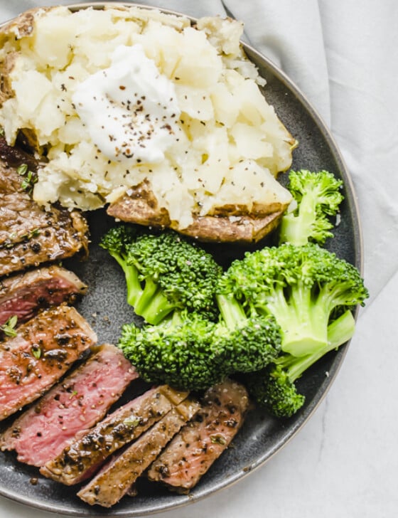 Seasoned cooked steak sliced on a plate with steamed broccoli and a baked potato.