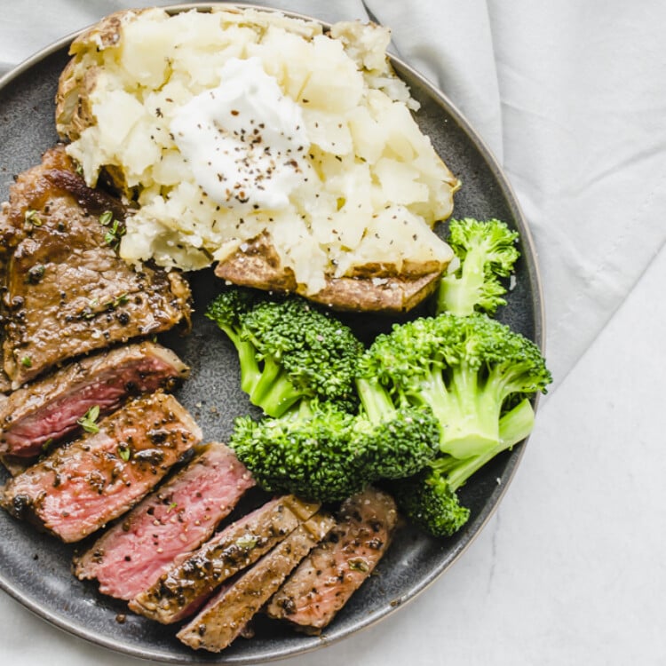 Seasoned cooked steak sliced on a plate with steamed broccoli and a baked potato.