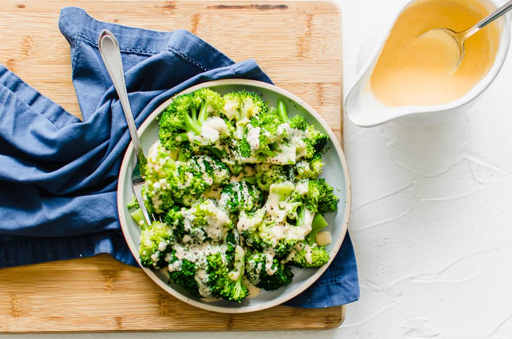 Steamed broccoli on a plate with homemade cheese sauce drizzled over top sitting on a wooden cutting board.