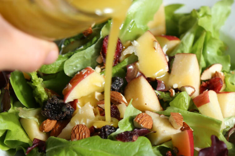 Homemade honey dijon vinaigrette being poured over a salad with chopped apples and almonds on top.
