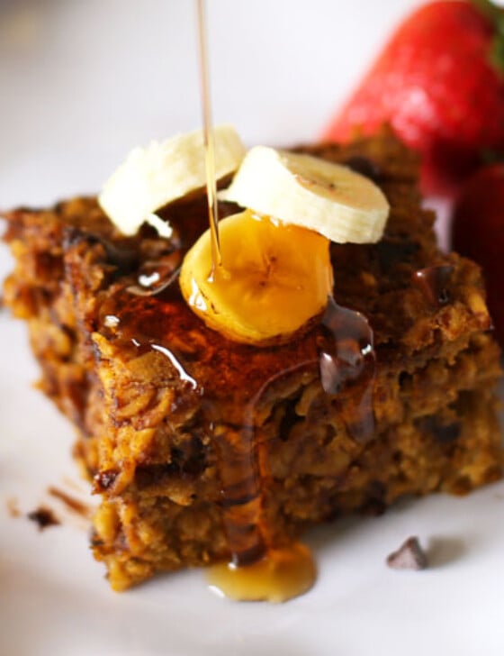 Slice of Pumpkin Baked Oatmeal on a white plate with banana slices and mini chocolate chips on top and maple syrup being poured on it.