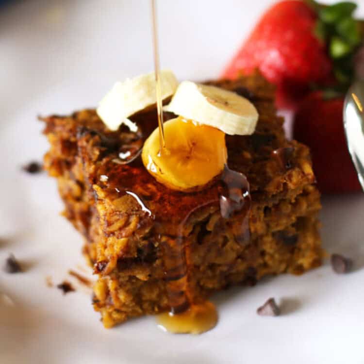 Slice of Pumpkin Baked Oatmeal on a white plate with banana slices and mini chocolate chips on top and maple syrup being poured on it.