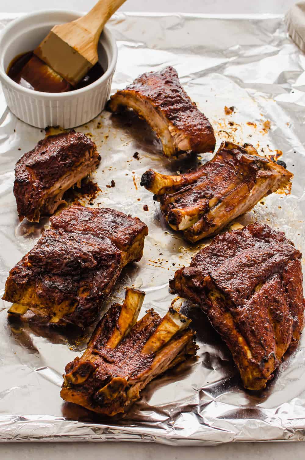 Crock pot baby back ribs that have been broiled on a foil-lined sheet pan with BBQ sauce on the side.