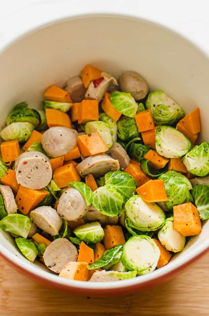 sliced Brussels sprouts, diced sweet potatoes, and sliced chicken sausage tossed in a white bowl with oil, salt, and pepper