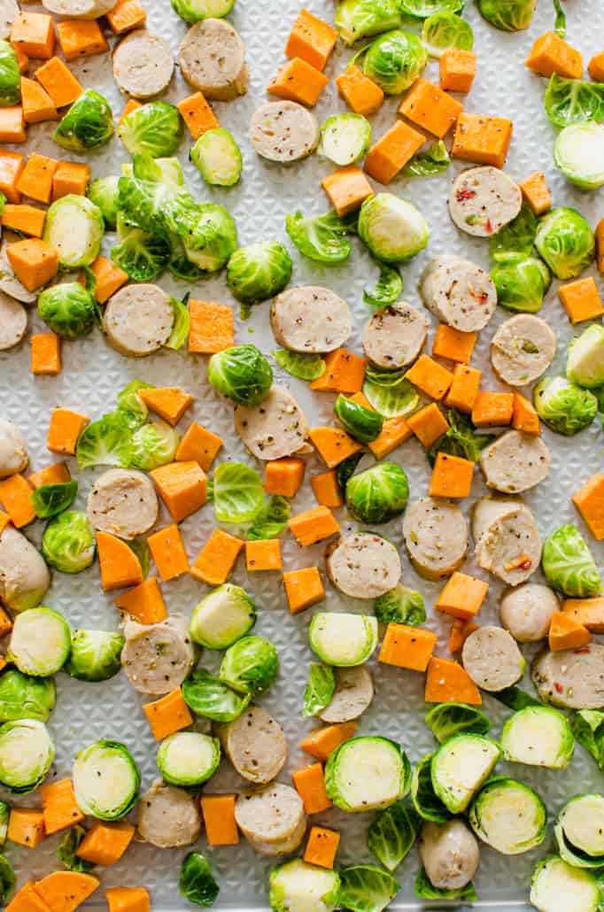 sliced Brussels sprouts, sweet potatoes, and chicken sausage spread out on a rimmed metal sheet pan before going in the oven