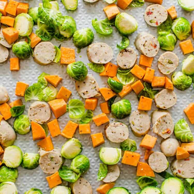 sliced Brussels sprouts, sweet potatoes, and chicken sausage spread out on a rimmed metal sheet pan before going in the oven