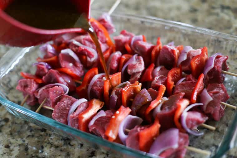 Raw beef kabobs in a baking dish with marinade being poured over them.