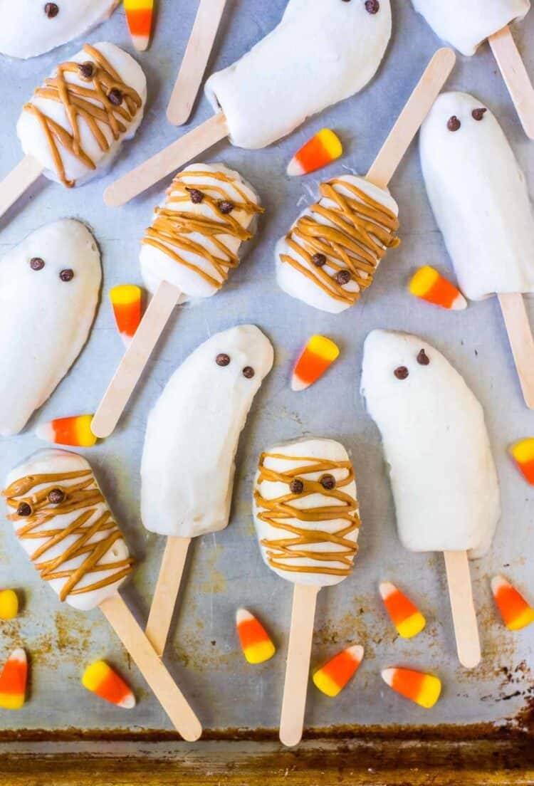 Halloween banana popsicles laid out - half bananas on a stick some with eyes to look like ghosts and others with peanut butter drizzle to look like mummies.