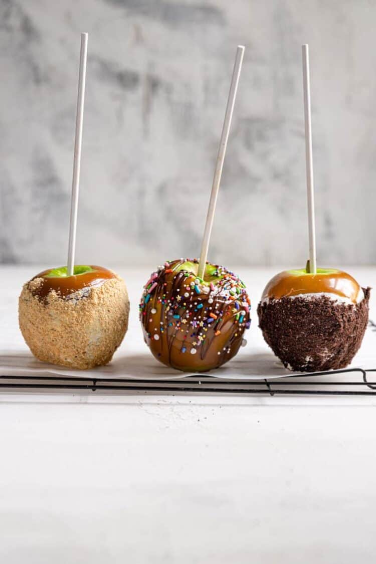 Three homemade caramel apples in a line, each with different toppings (crushed graham crackers, drizzled chocolate and sprinkles, crushed chocolate cookies).